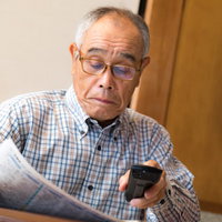 テレビのリモコンを持ちながら新聞を読むシニアの写真