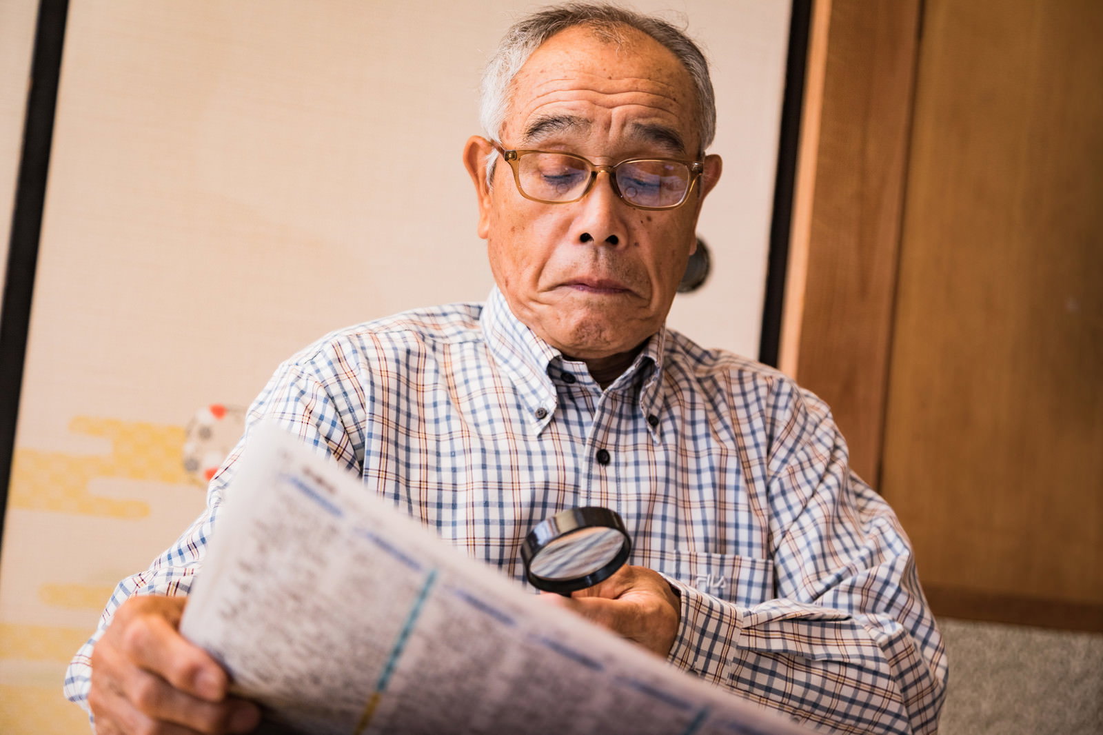 拡大鏡を使って新聞を読むチェック柄シャツの高齢男性［モデル：まーちゃん］