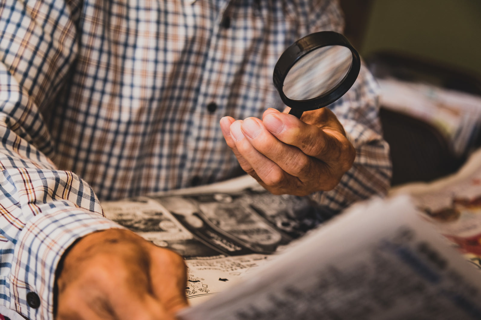 チェック柄のシャツを着た男性が拡大鏡を使って新聞を読んでいる様子［モデル：まーちゃん］