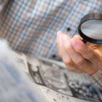 チェック柄のシャツ姿で手持ち拡大鏡を使って新聞を読む高齢男性の手の写真