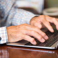 チェック柄シャツを着た高齢者が慣れた手つきでキーボードをタイピングする手元の写真