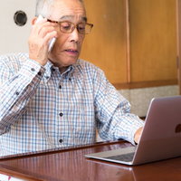 電話しながらパソコンを操作するシニア男性の写真