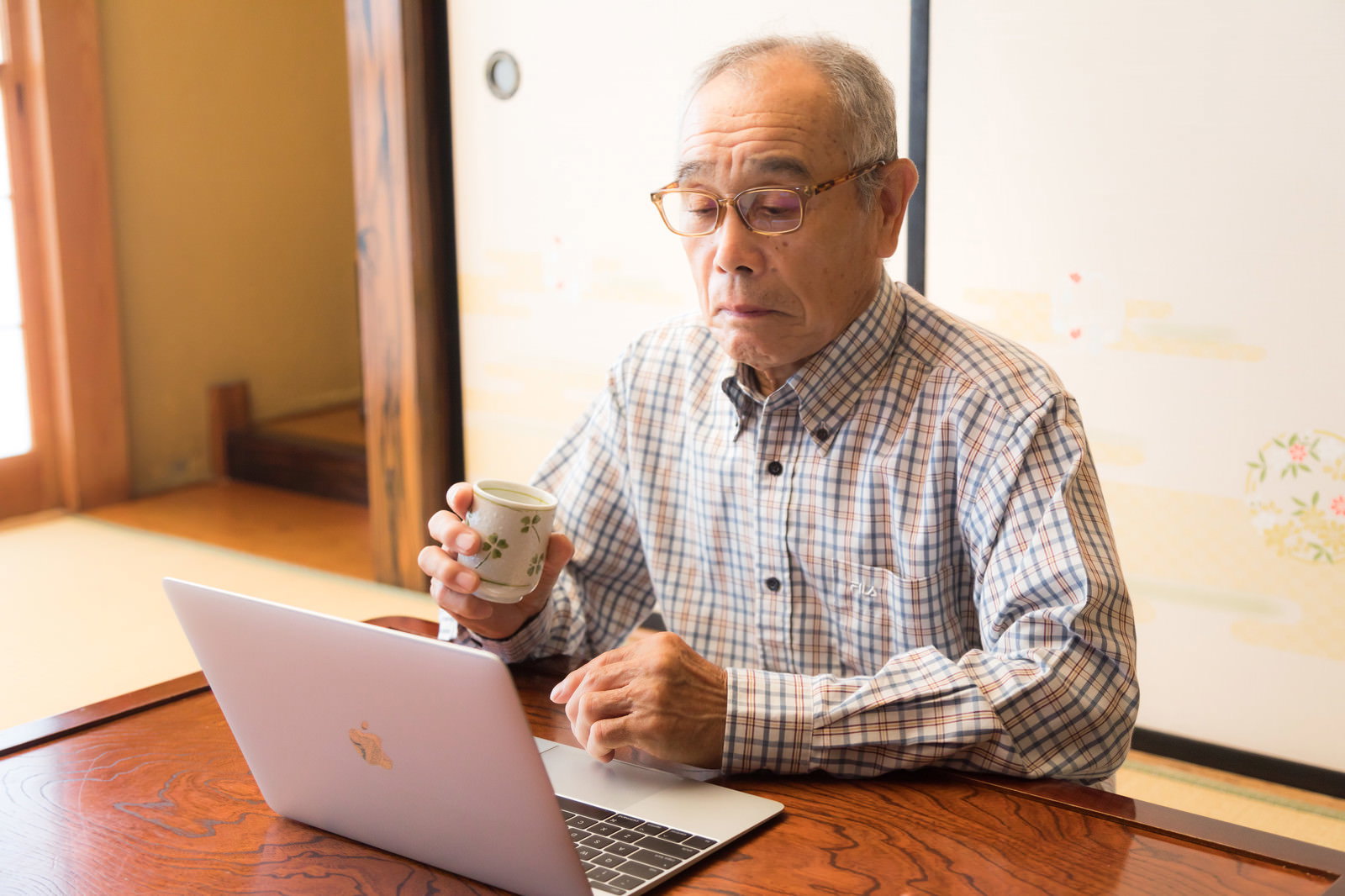 眼鏡をかけたおじいさんがノートパソコンを操作しながら湯呑を持っている様子［モデル：まーちゃん］