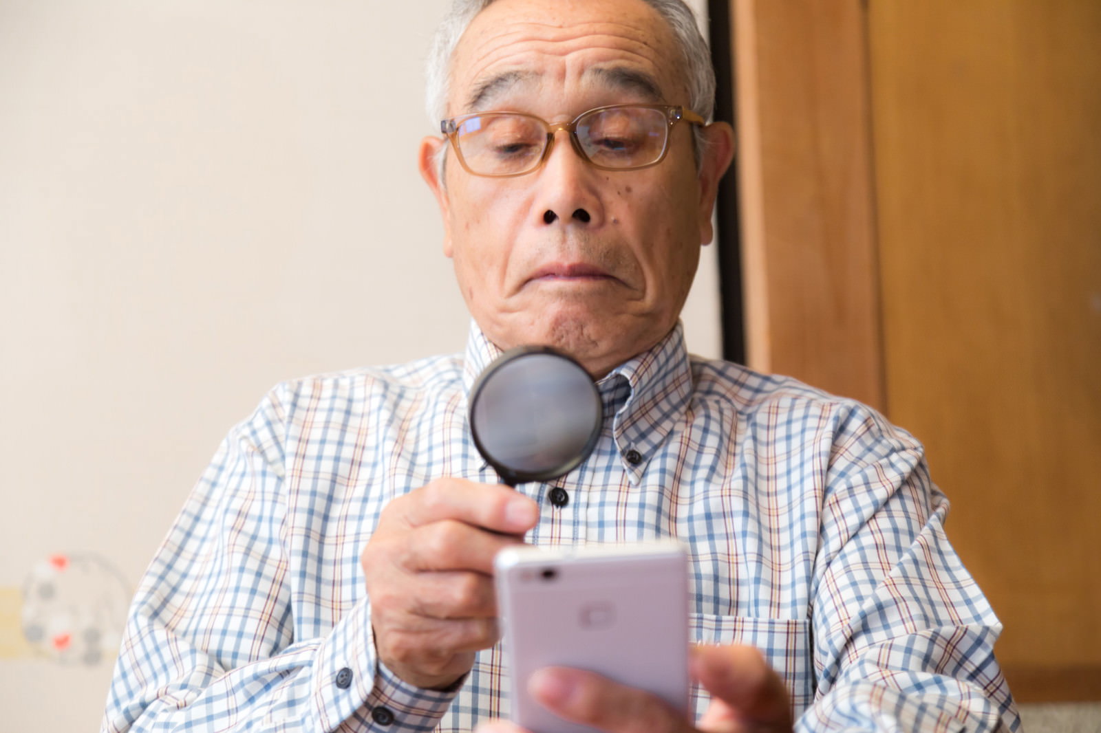 拡大鏡を使ってスマートフォンの画面を見ている眼鏡をかけた高齢男性［モデル：まーちゃん］