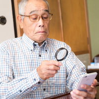 ガラケーの小さい文字を拡大鏡で確認する高齢男性の写真