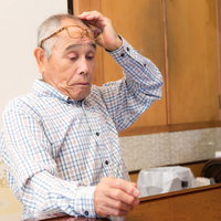 眼鏡をかけて机の上で探す高齢者男性、困り顔でデスクワークの写真