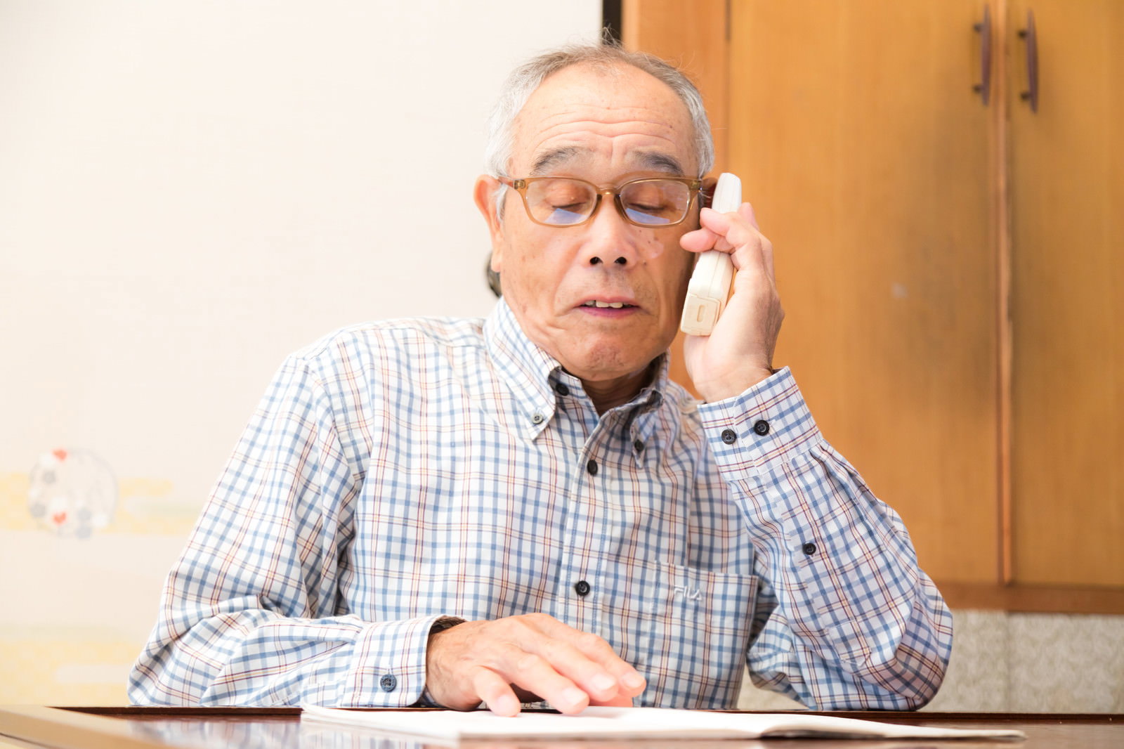 眼鏡をかけた高齢男性が通販カタログを見ながら電話で注文している［モデル：まーちゃん］