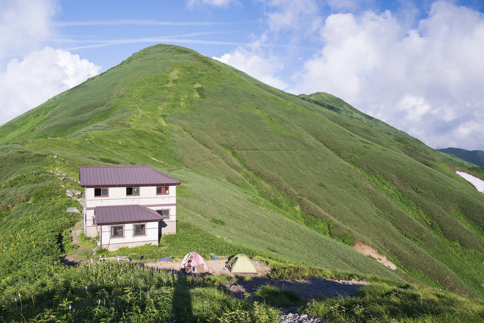 緑の山々の中腹に建つ白い梅花皮小屋と青空が広がる山岳風景