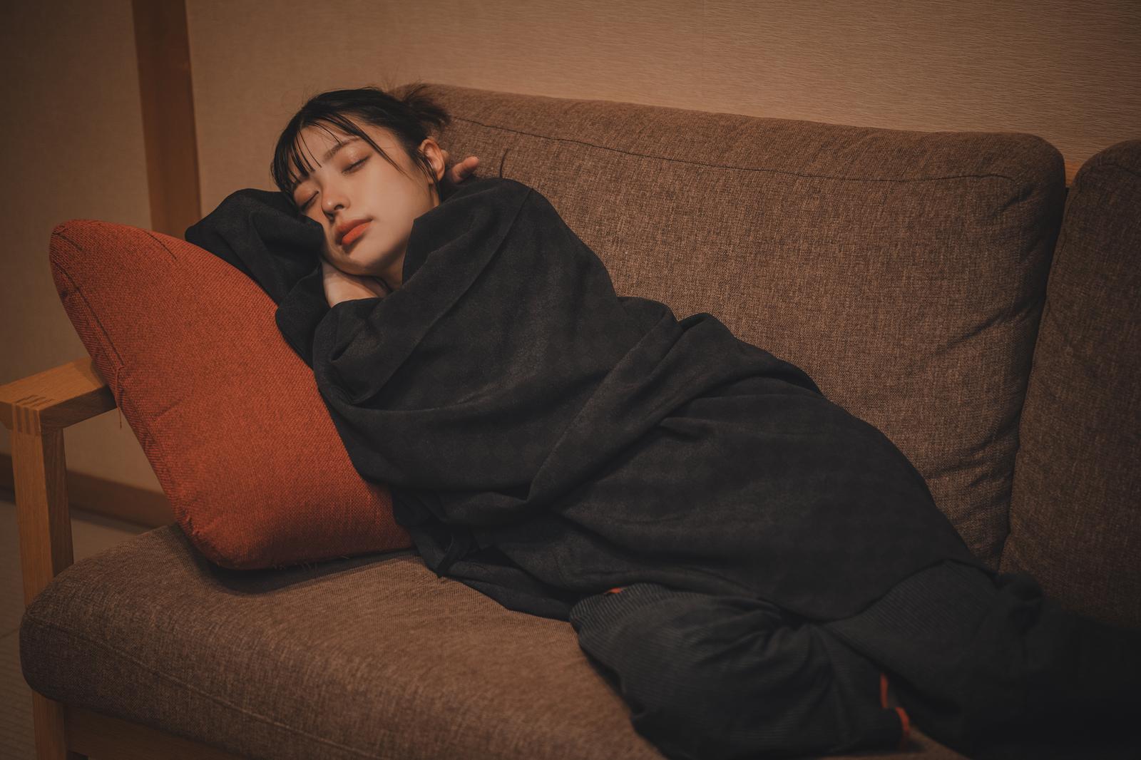 黒いパーカーを着てソファーで眠る女性。赤いクッションを枕にしている。［モデル：高尾実生］