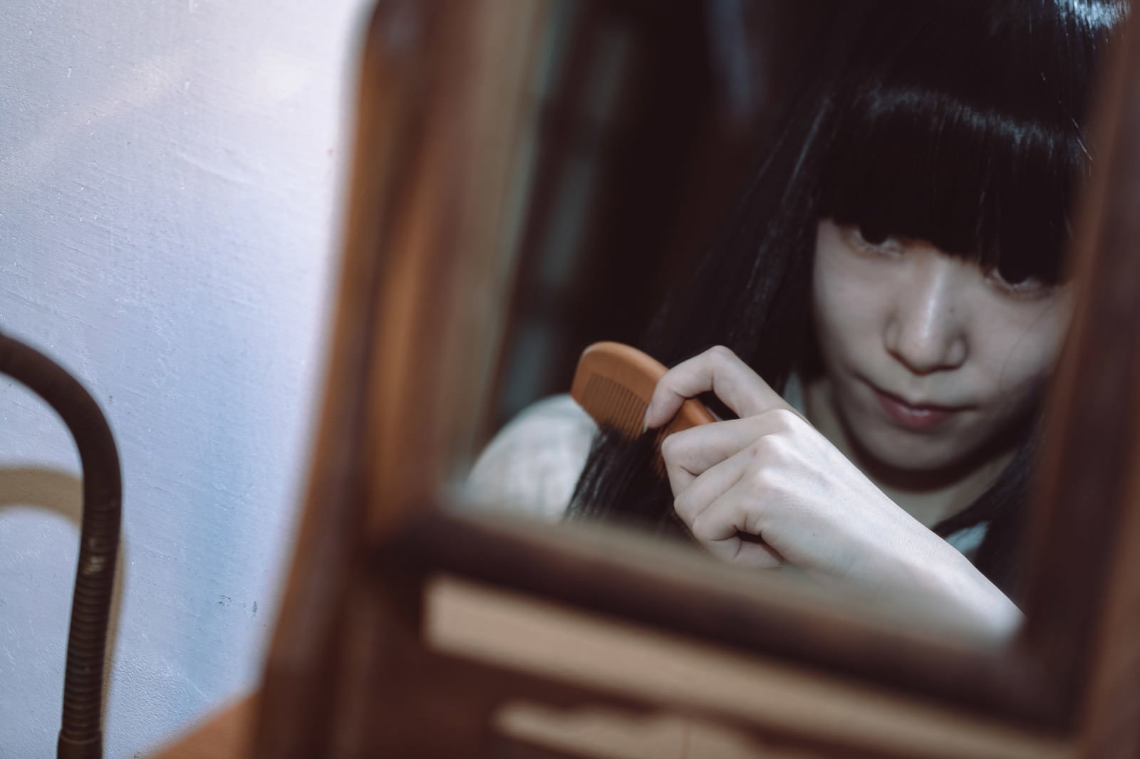 薄暗い部屋で鏡を見ながら櫛で髪を梳かす前髪パッツンの女性［モデル：緋真煉］