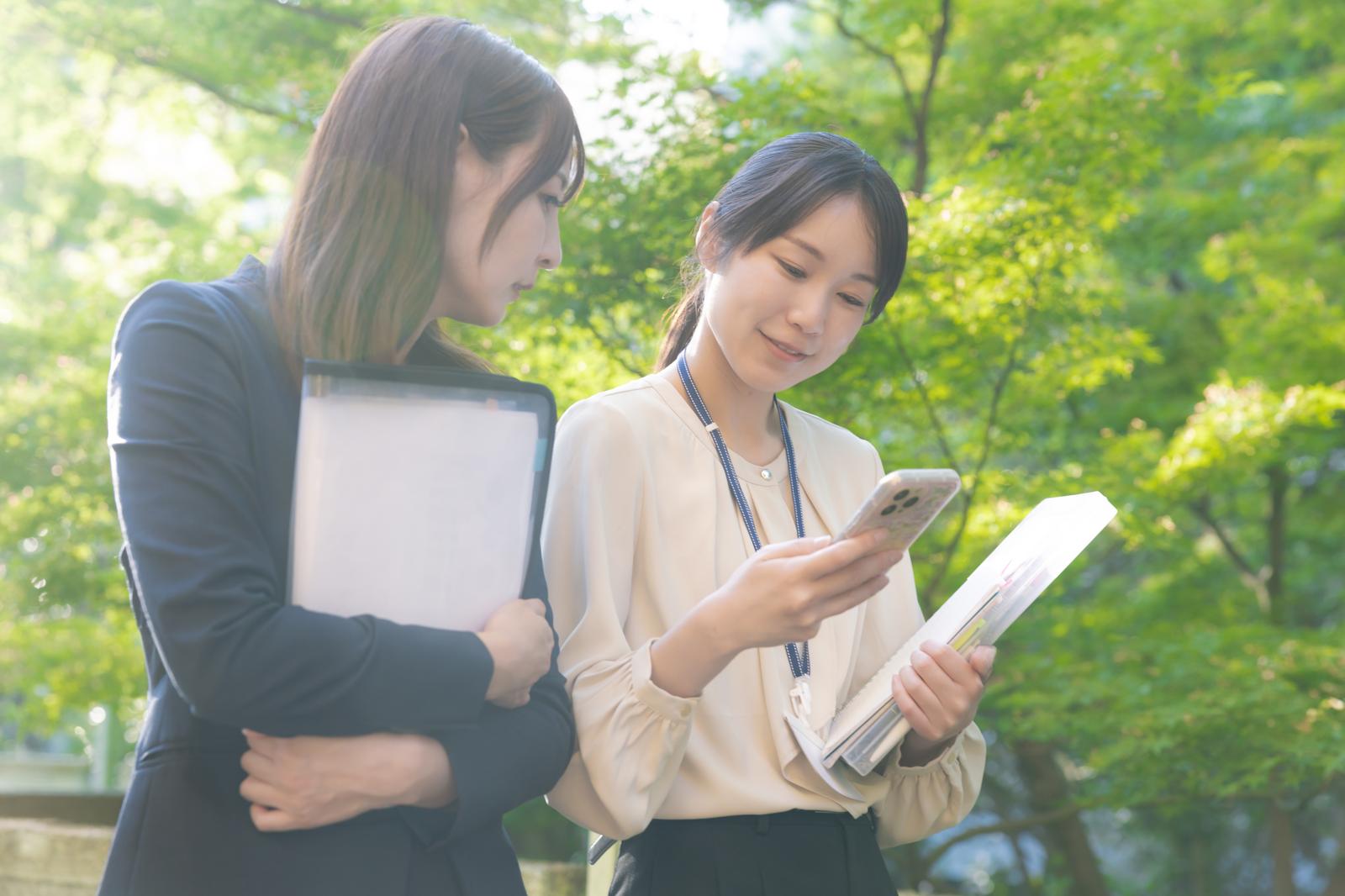 屋外でスマホと資料を確認する女性社員2人［モデル：SAKI あゆみん］