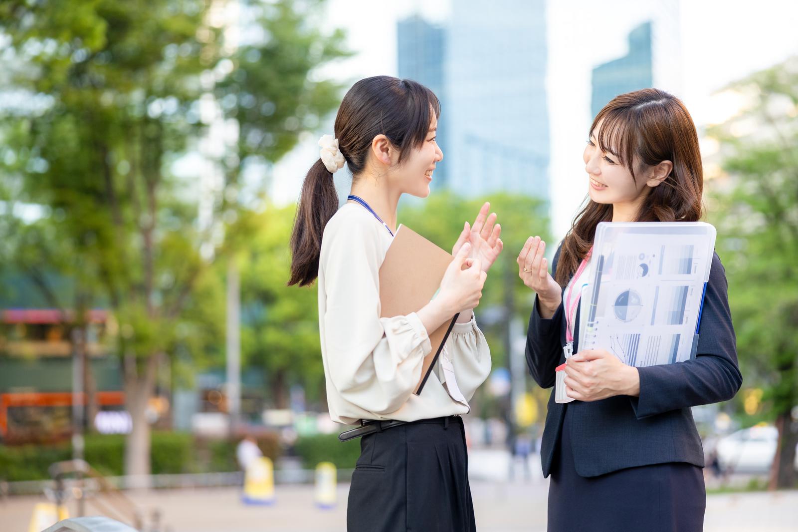 資料を持った2人の女性社員がビルと新緑を背景に立ち話をしている［モデル：SAKI あゆみん］
