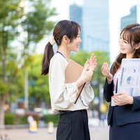 資料をもって立ち話をする女性社員ふたり、ビジネス打ち合わせの写真