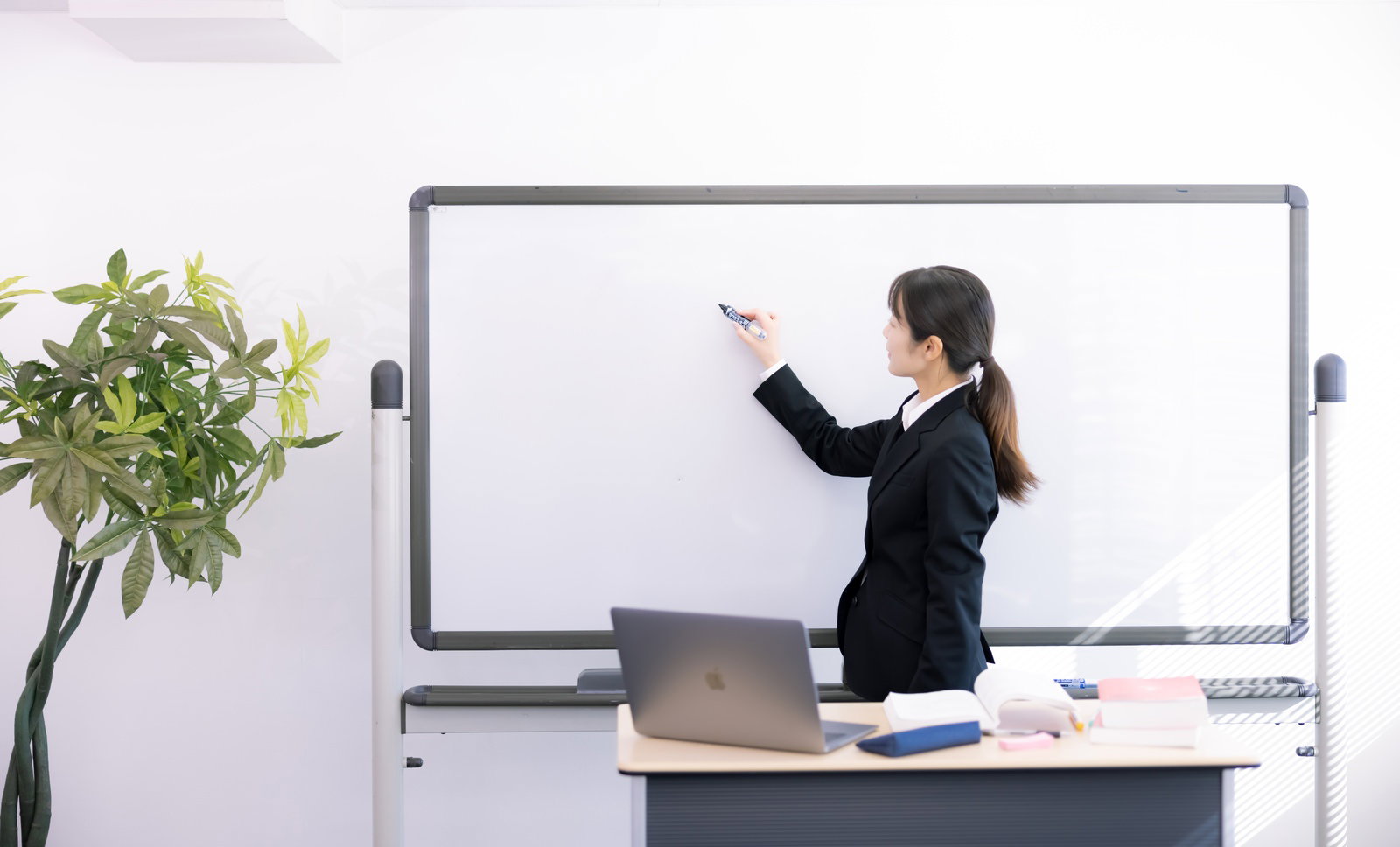 ホワイトボードの前でマーカーを持って説明する黒いスーツの女性講師［モデル：SAKI］