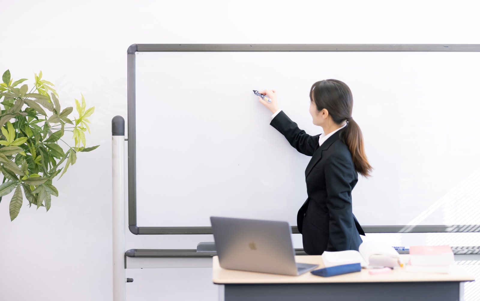 ホワイトボードに書き込みながら説明する黒いスーツの女性講師［モデル：SAKI］