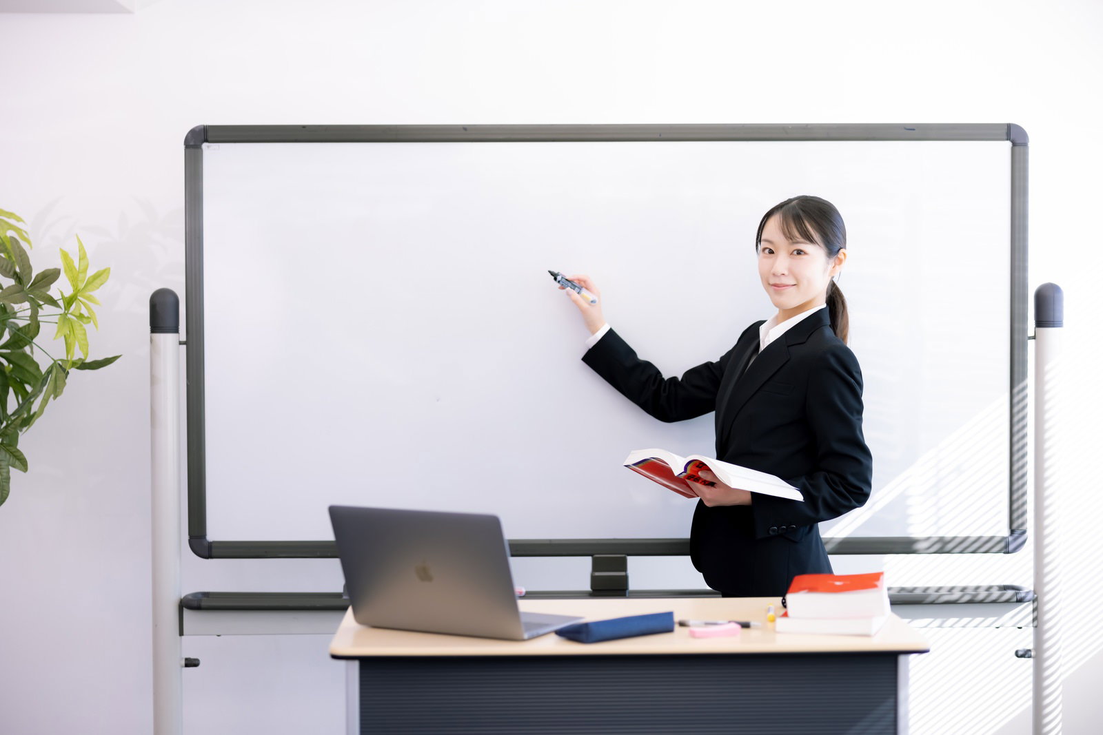 ホワイトボードの前で振り返って指導する黒いスーツの女性講師［モデル：SAKI］