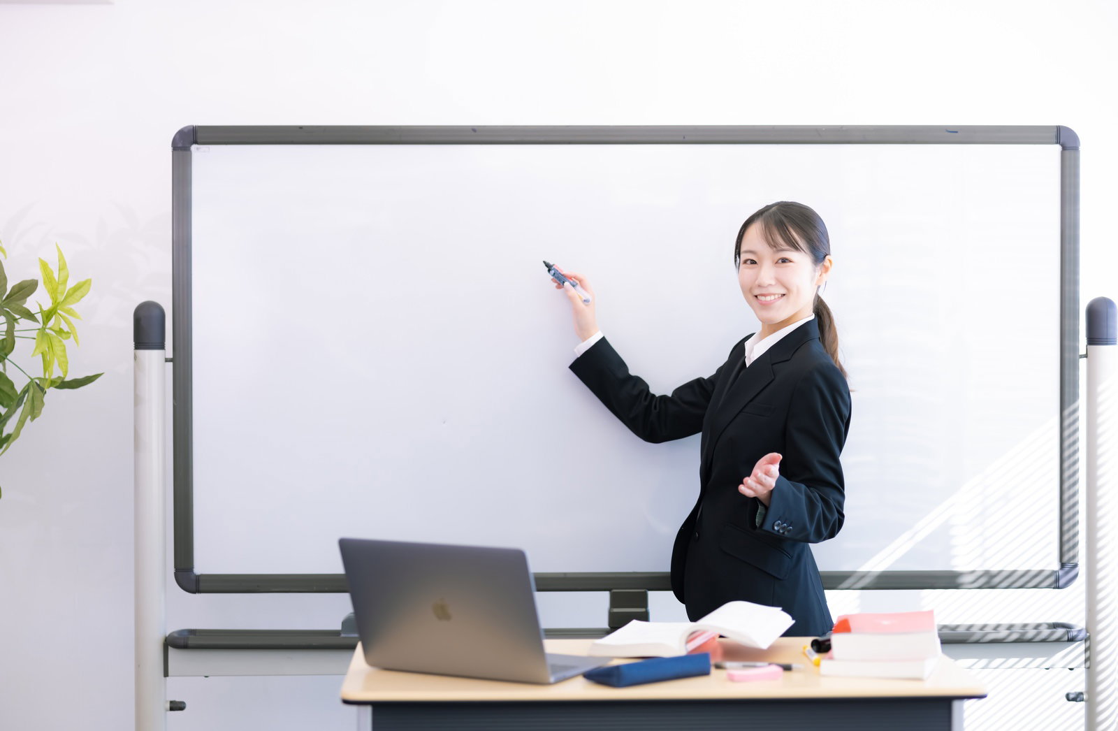 黒いスーツを着た女性がホワイトボードの前で指し棒を持ち笑顔でプレゼンテーション［モデル：SAKI］
