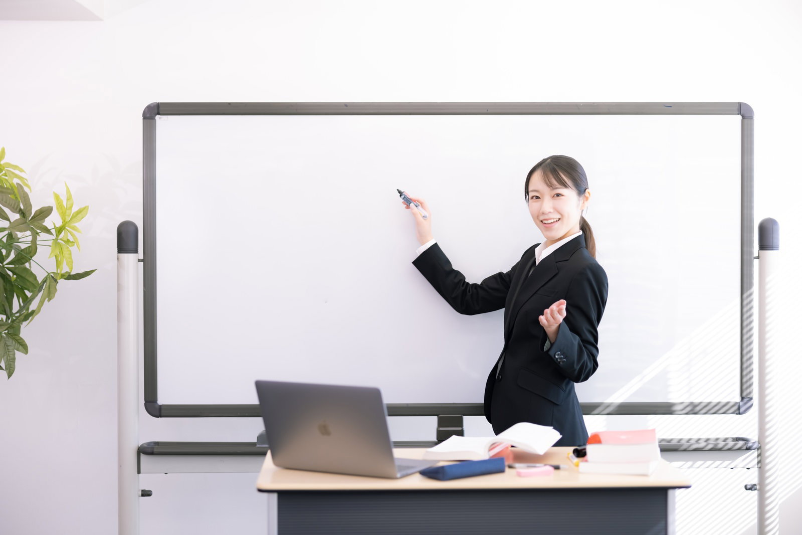 ホワイトボードの前で指し棒を持ち説明している黒いスーツの女性講師［モデル：SAKI］