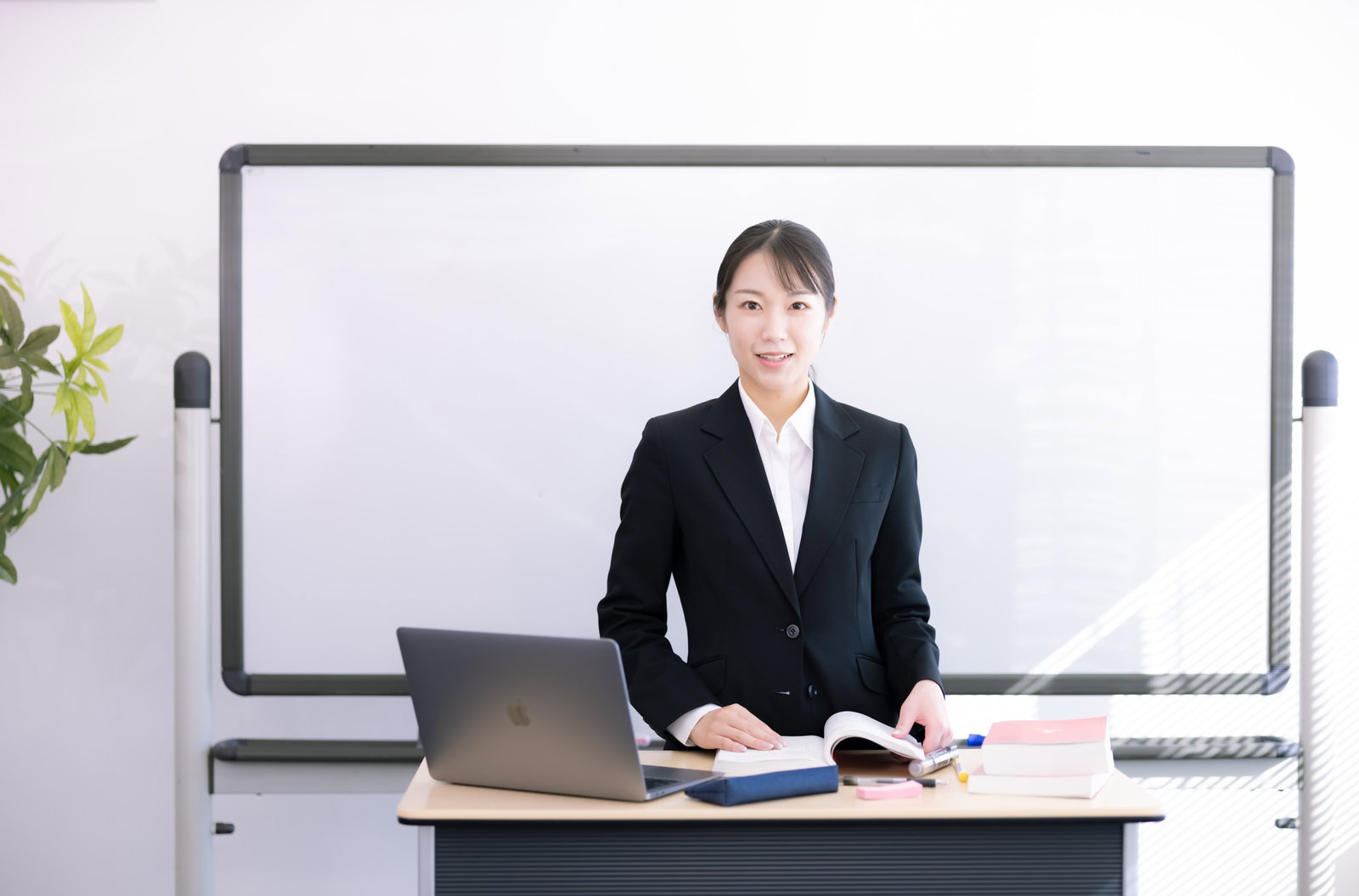 黒いスーツを着た女性講師がホワイトボードの前で授業を行っている様子［モデル：SAKI］
