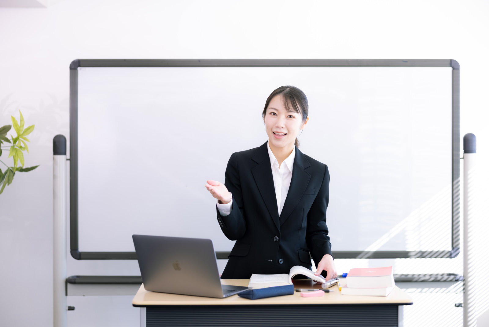 ホワイトボードの前で自己紹介する黒いスーツの女性講師［モデル：SAKI］