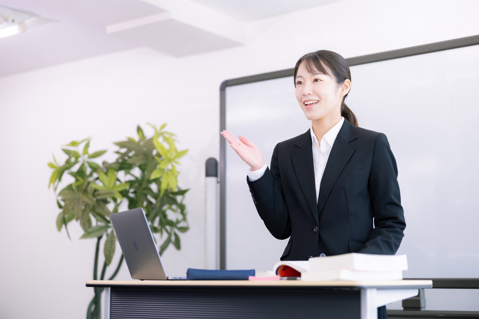 ホワイトボード前で手を上げながらプレゼンする黒スーツの女性講師［モデル：SAKI］