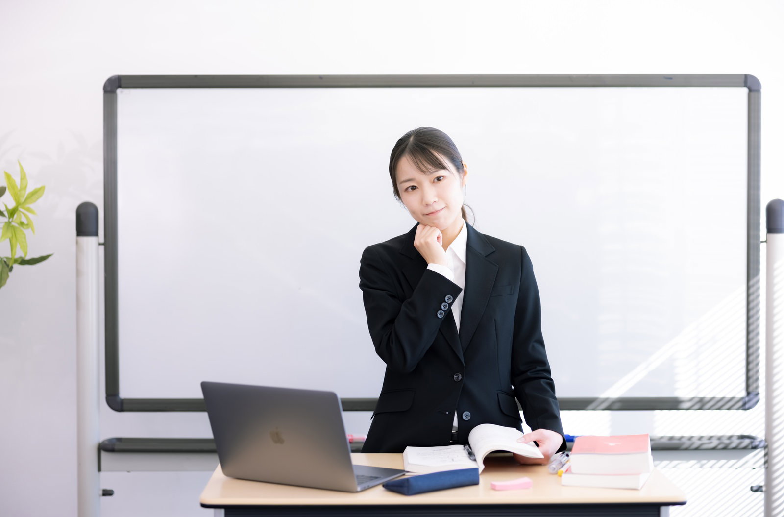 ホワイトボードの前で困惑した表情を見せる黒いスーツの女性講師［モデル：SAKI］