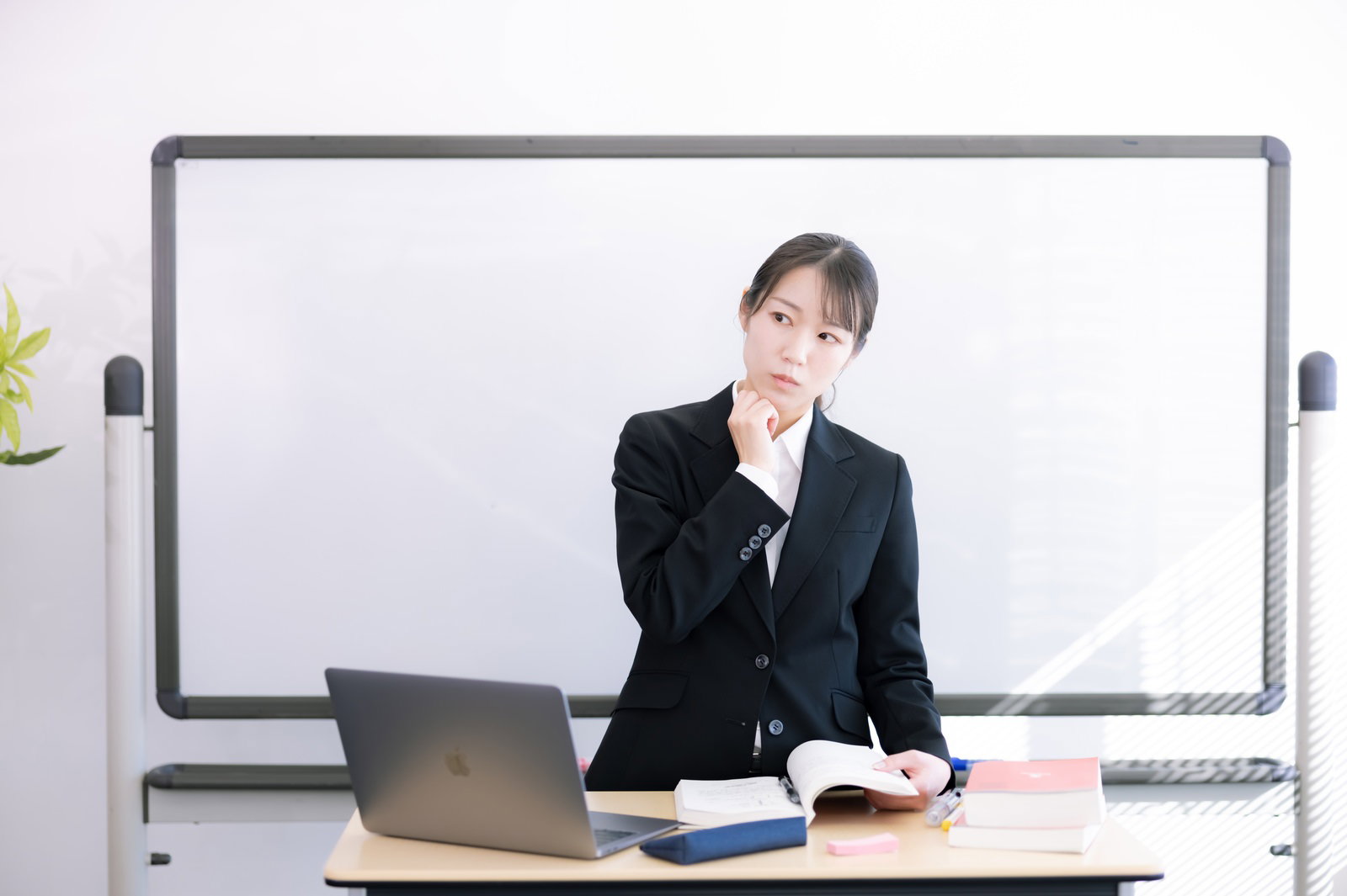 ホワイトボード前で困った表情を見せる黒いスーツ姿の女性講師［モデル：SAKI］