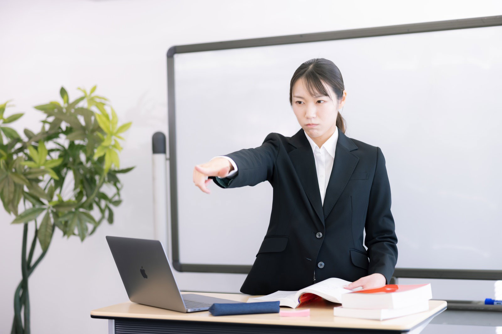ホワイトボード前で指を指しながら注意を促す黒いスーツの女性講師［モデル：SAKI］