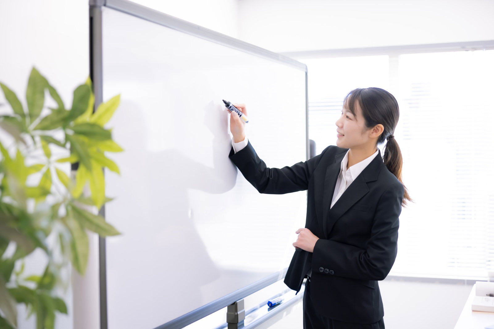 黒いスーツを着た女性講師がホワイトボードで説明している様子［モデル：SAKI］