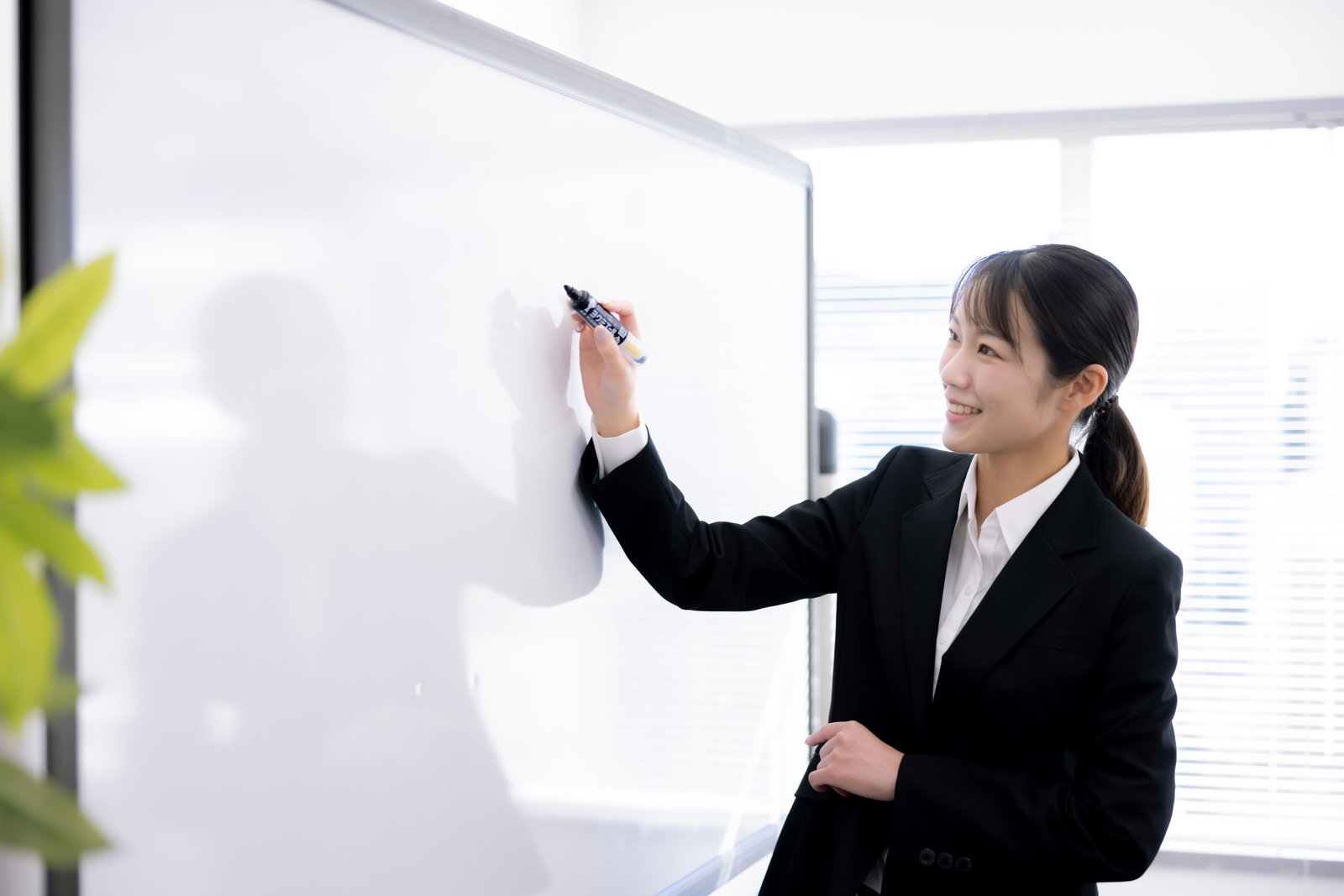 黒いスーツを着た女性講師がホワイトボードの前で笑顔で講演している様子［モデル：SAKI］