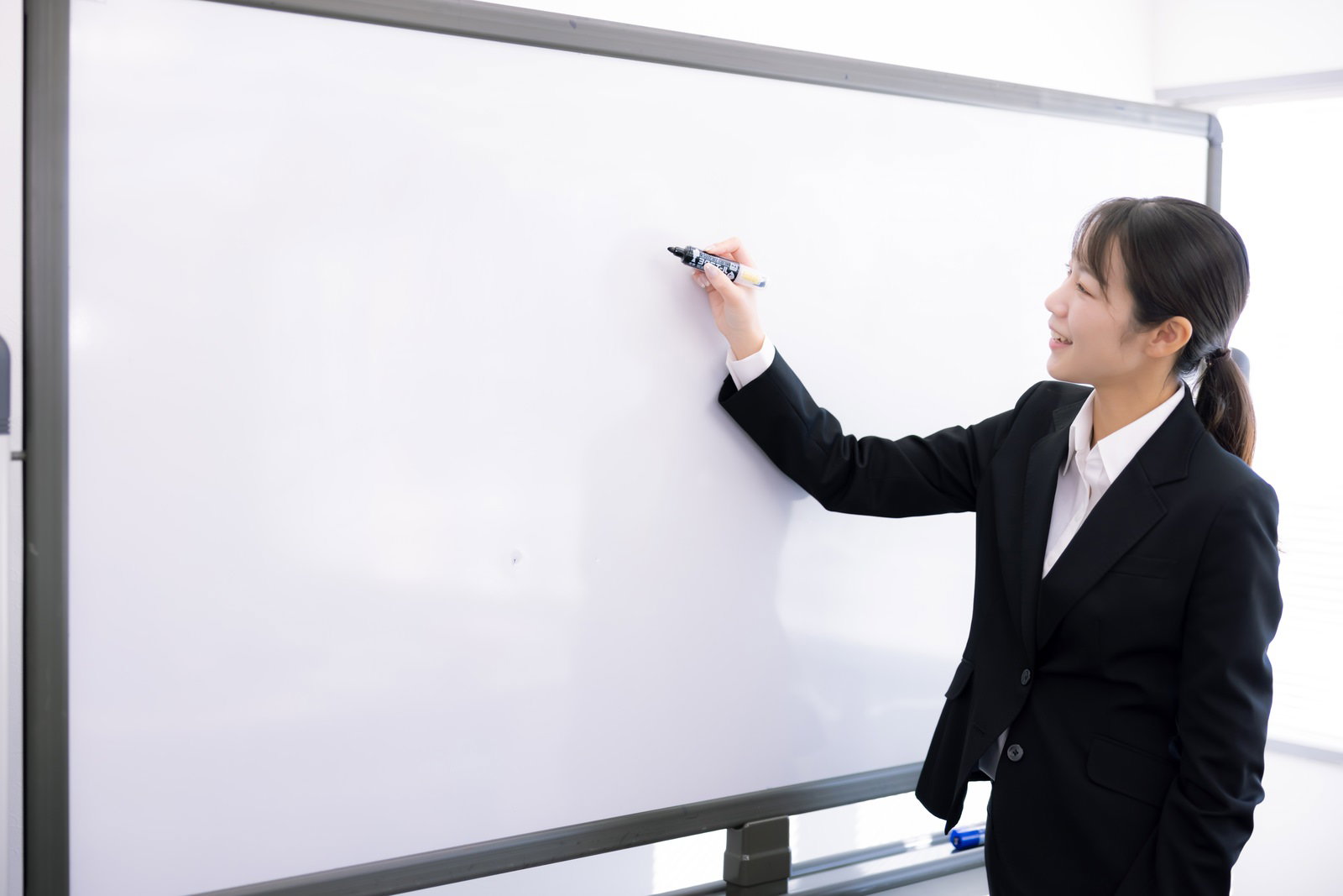 ホワイトボードにペンで書きながら説明する黒いスーツの女性講師［モデル：SAKI］