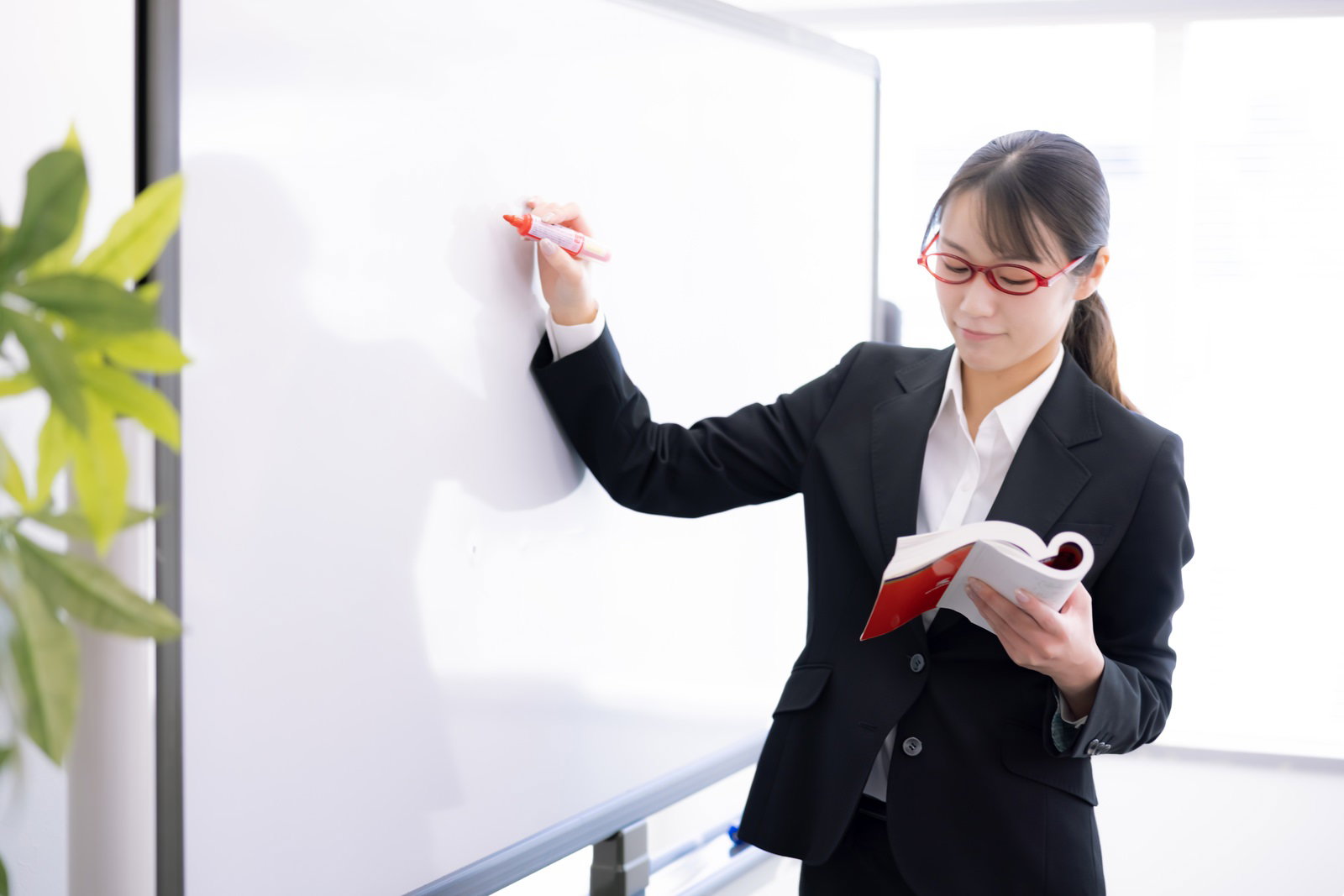 ホワイトボードにマーカーで書き込みをする黒いスーツ姿の女性講師［モデル：SAKI］