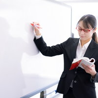 黒いスーツの女性講師がホワイトボードで授業をする様子の写真