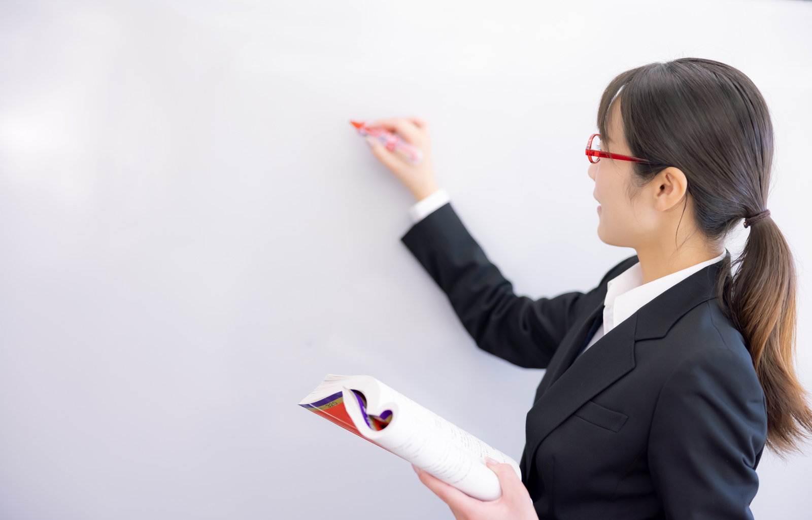 黒いスーツを着た女性講師がマーカーでホワイトボードに記入している様子［モデル：SAKI］