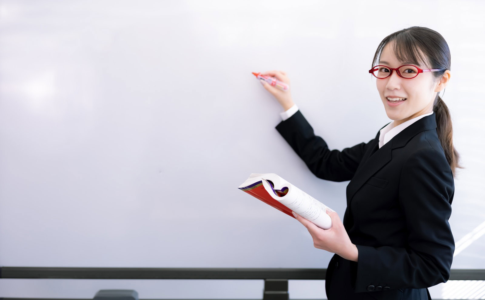 ホワイトボードの前で赤いマーカーを持って説明する黒いスーツ姿の女性講師［モデル：SAKI］