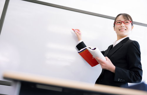 赤い眼鏡をかけた女性講師がホワイトボードの前で説明