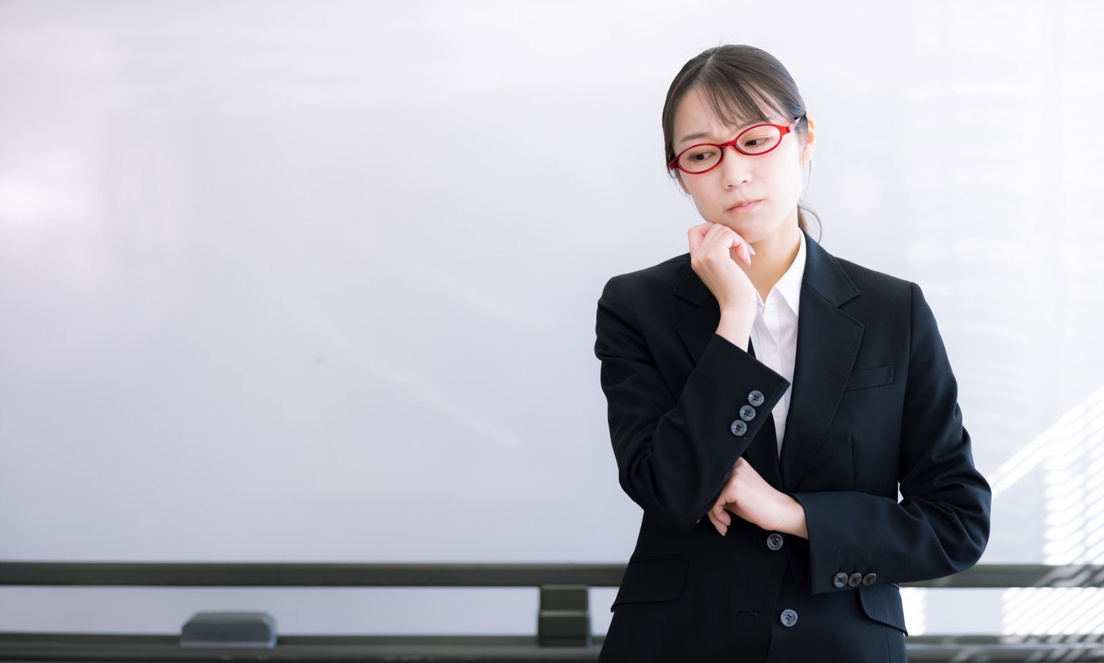 赤い眼鏡をかけた女性講師がホワイトボードの前で困惑している様子［モデル：SAKI］