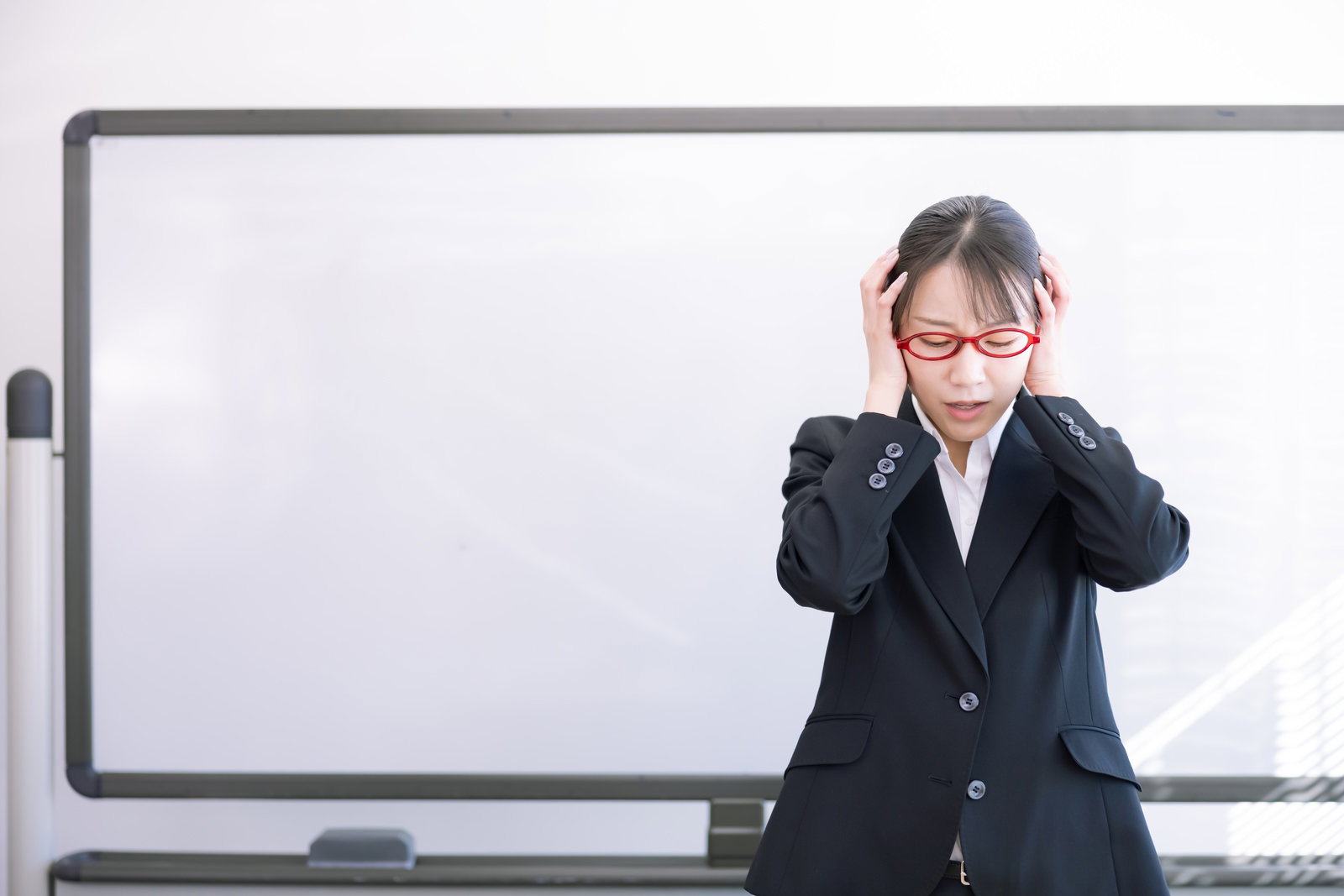 ホワイトボードの前で頭を抱える赤い眼鏡をかけた黒いスーツの女性講師［モデル：SAKI］