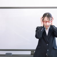 ホワイトボード前で頭を抱える赤い眼鏡の女性講師の写真
