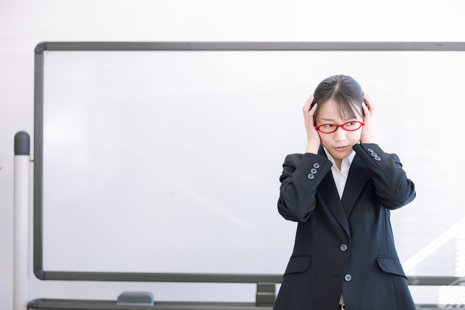 ホワイトボード前で両手で頭を抱えて後悔する赤い眼鏡をかけた女性講師［モデル：SAKI］