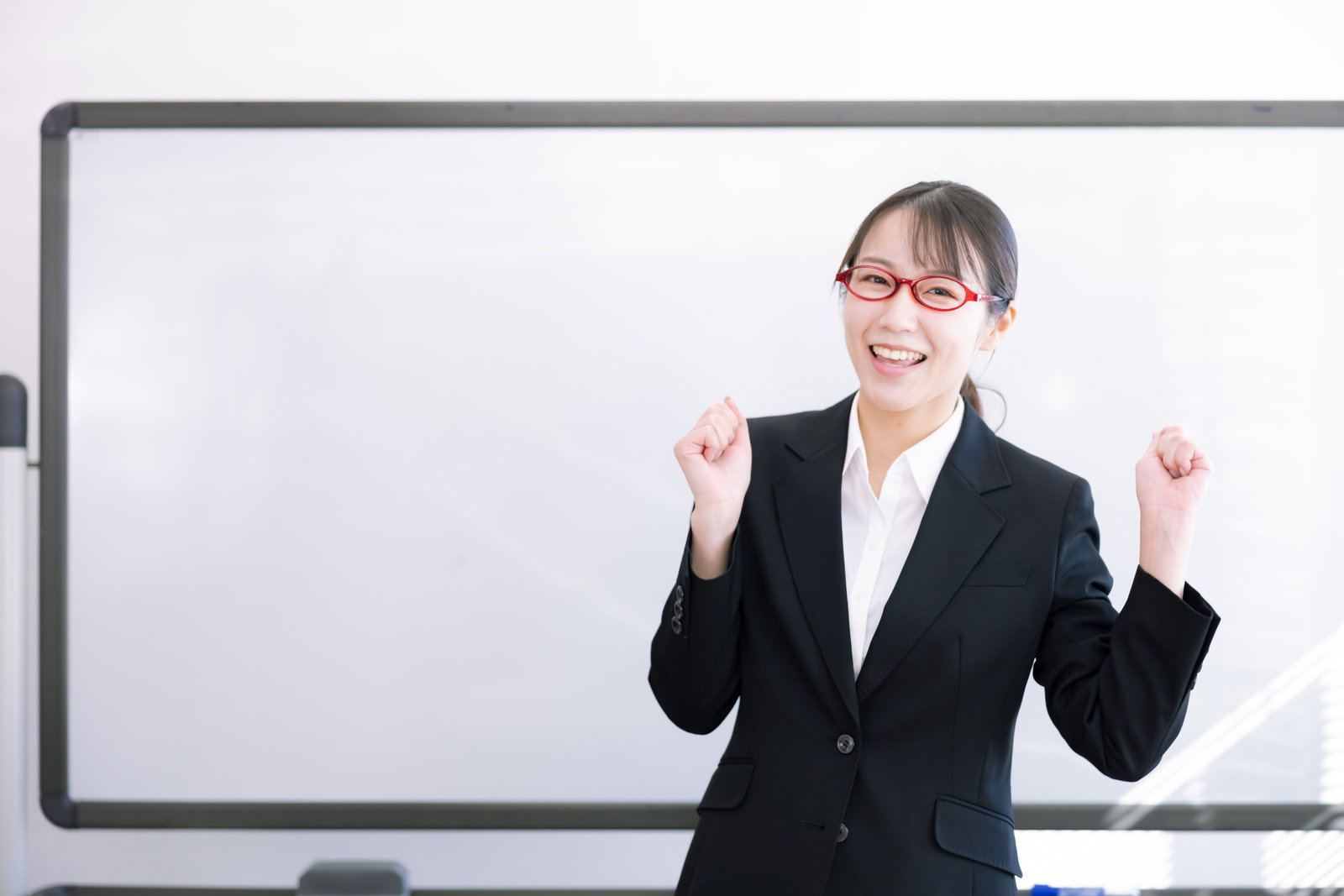 ガッツポーズをする笑顔の女性講師がホワイトボード前に立つ［モデル：SAKI］