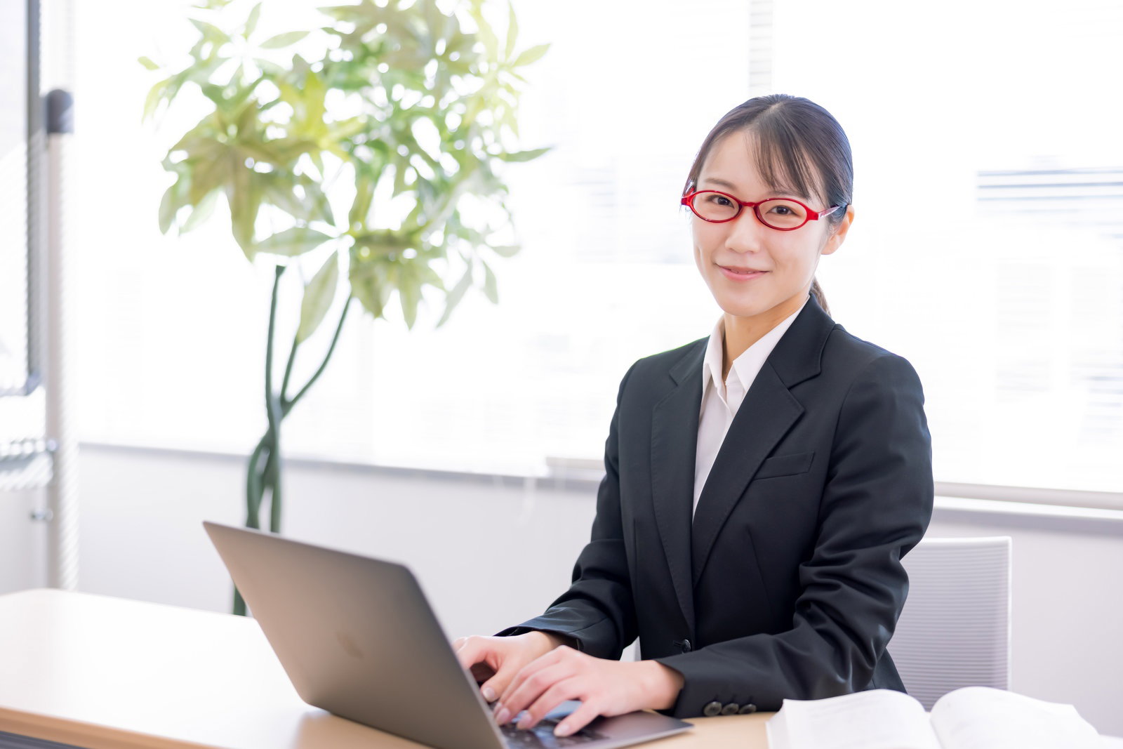 赤い眼鏡をかけたスーツ姿の女性がデスクでノートパソコンを操作している［モデル：SAKI］