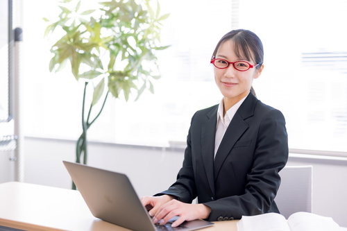 赤い眼鏡をかけた秘書課の女性がデスクでノートパソコンを操作