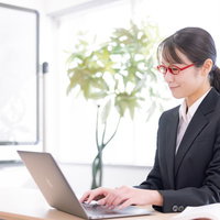 デスクワークしながらランチに何を食べようか考える黒いスーツの女性社員の写真