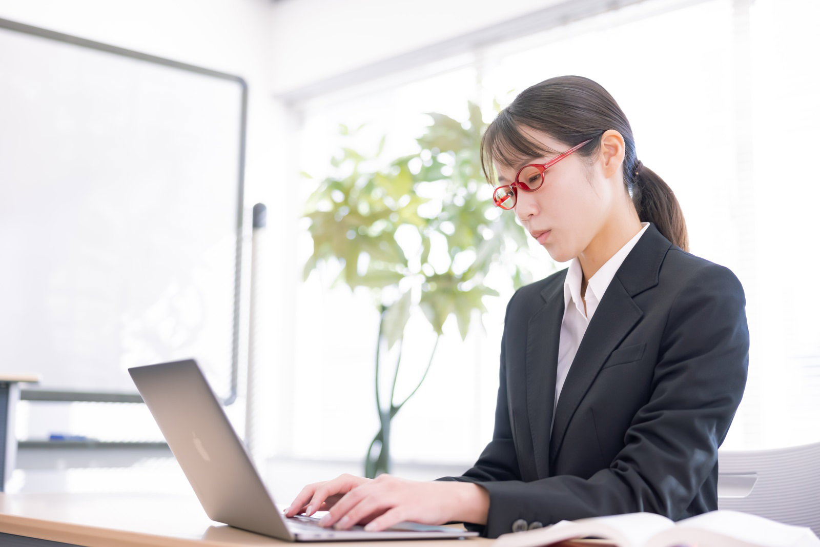 オフィスのデスクでノートパソコンに向かう黒いスーツの女性社員［モデル：SAKI］