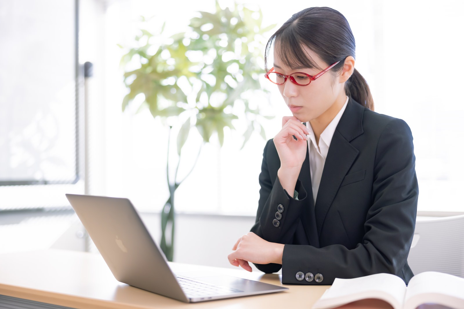 スーツを着た女性がデスクでノートパソコンを見ながら首をかしげている様子［モデル：SAKI］