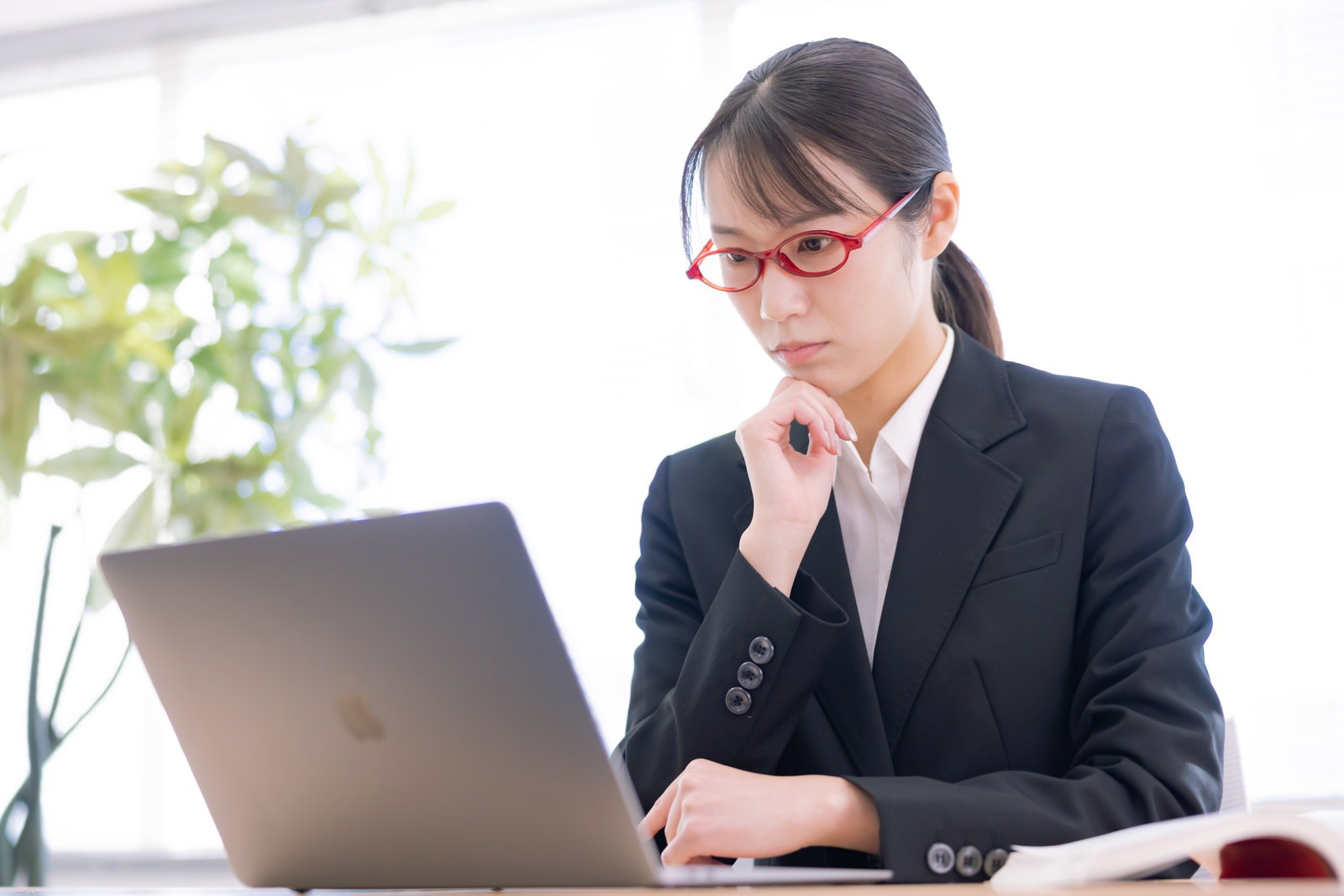 スーツを着た女性がデスクでパソコンに向かい手を顎に当てて考えている［モデル：SAKI］