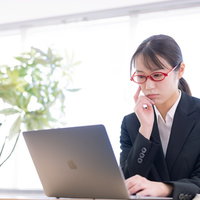 ノートパソコンの画面を見つめて考え込むスーツ姿の女性の写真