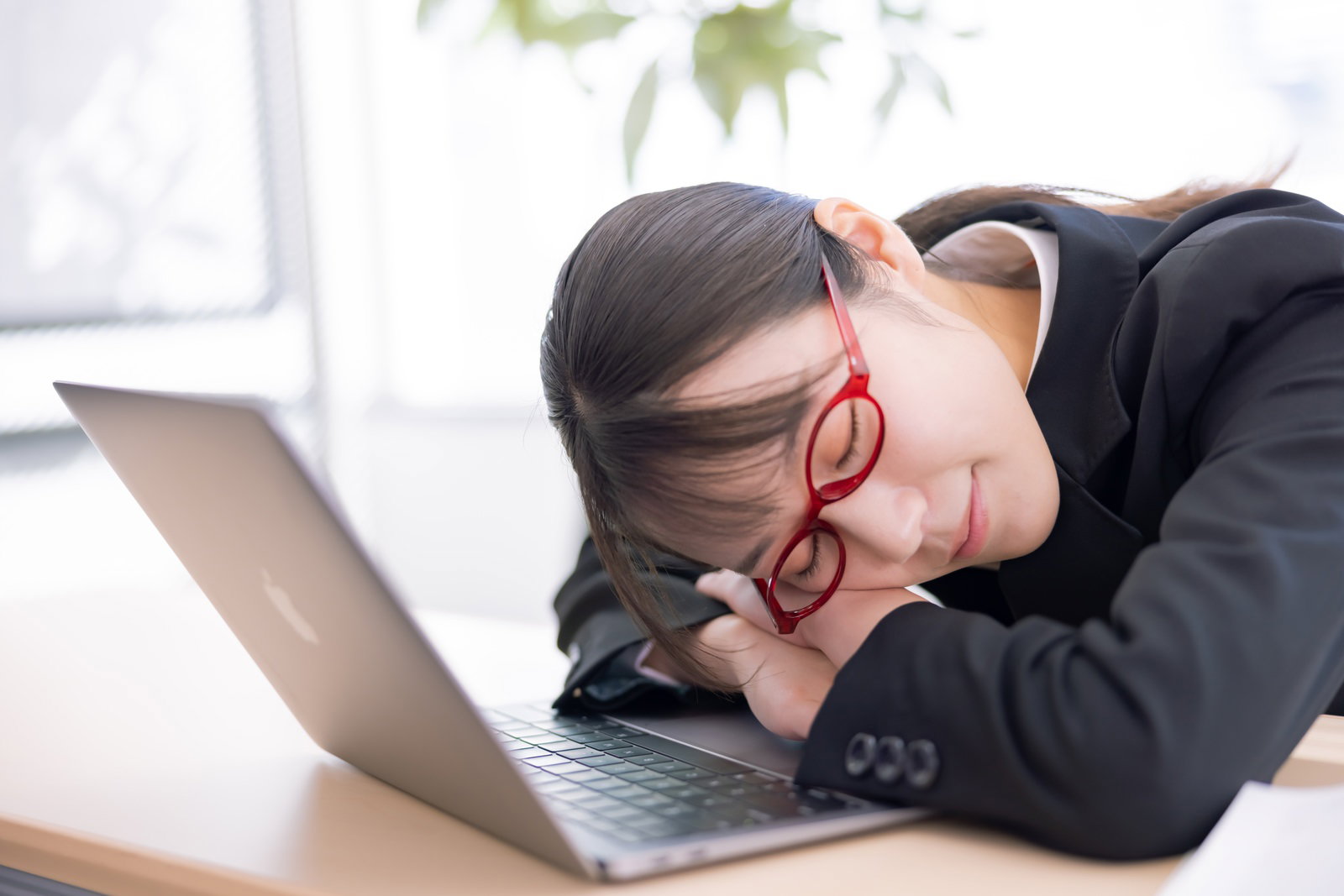 眼鏡をかけた女性がノートパソコンの前で机にうつ伏せになって眠っている［モデル：SAKI］
