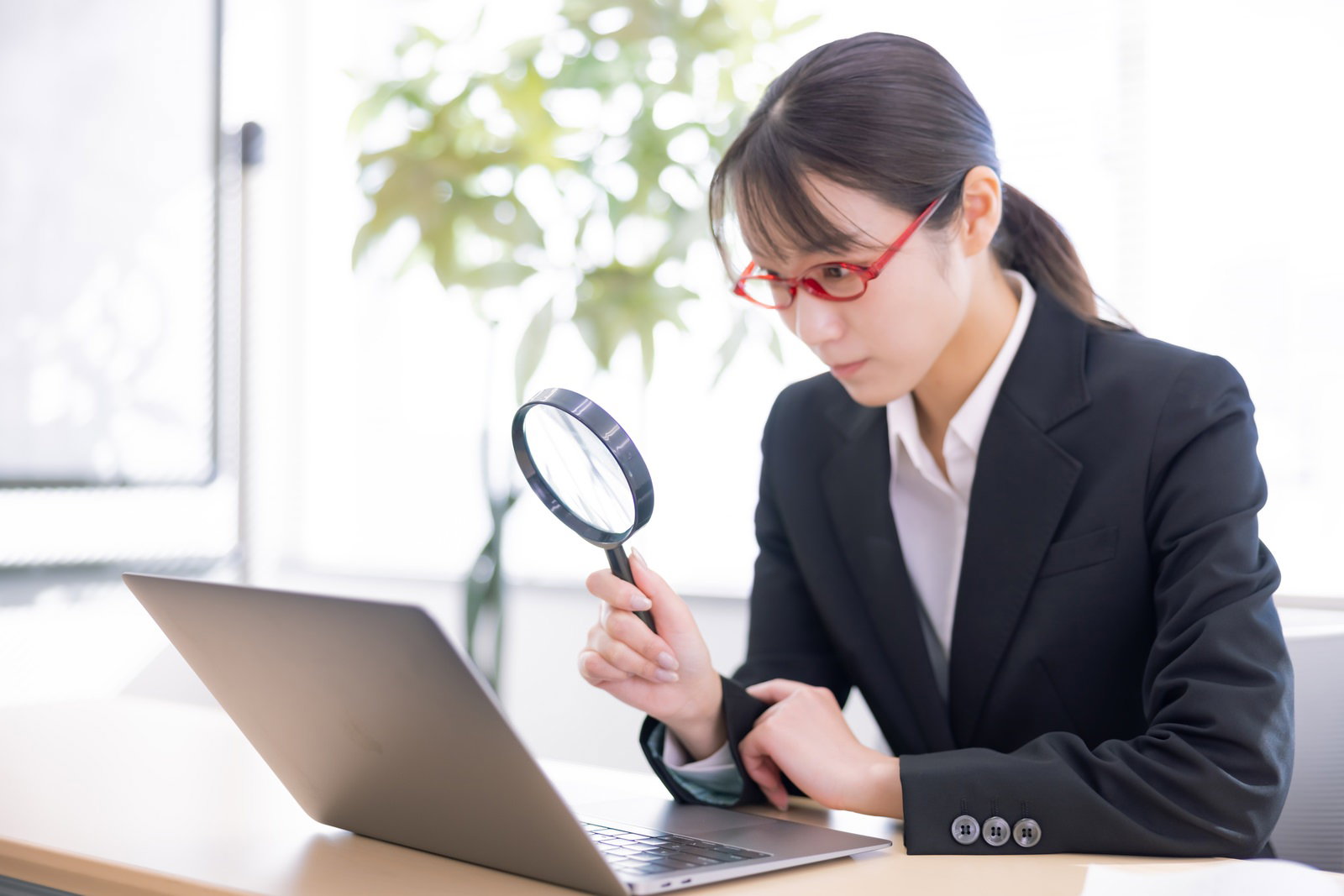 拡大鏡を使ってノートパソコンの画面をチェックするスーツ姿の女性社員［モデル：SAKI］